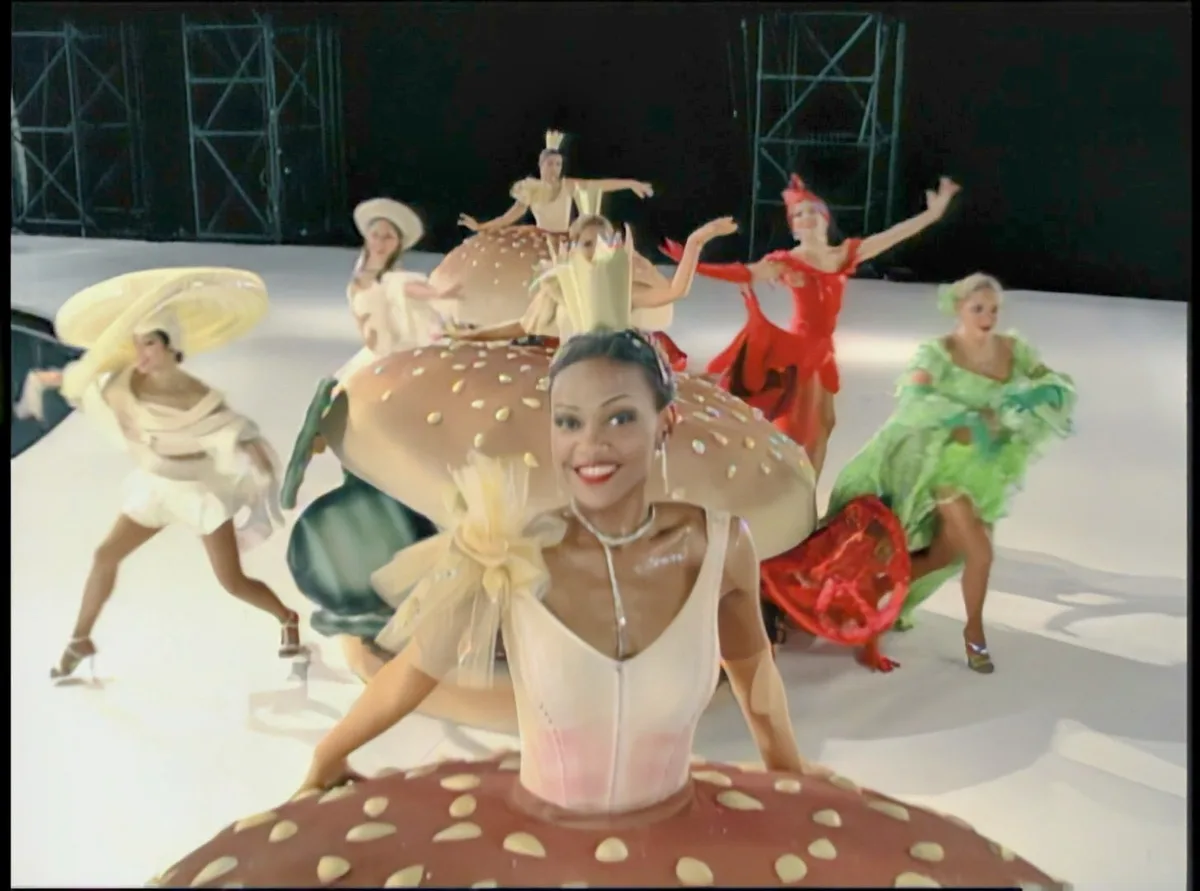 A woman in a white dress smiles while sitting atop a giant burger costume, surrounded by other dancers in colorful…