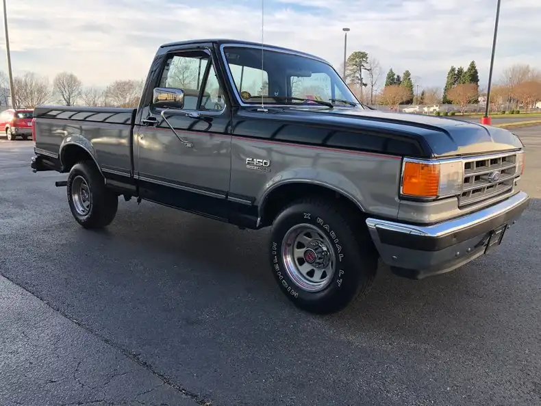 A silver and black 1980s Ford F-150 pickup truck is parked in an asphalt lot, featuring chrome bumpers, a black hood stripe…