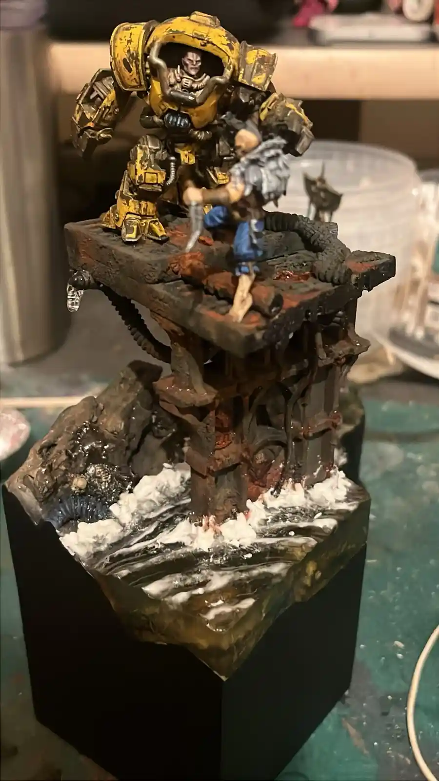 A painted miniature diorama shows a large yellow armored mech towering over a smaller human-like figure on a crumbling…