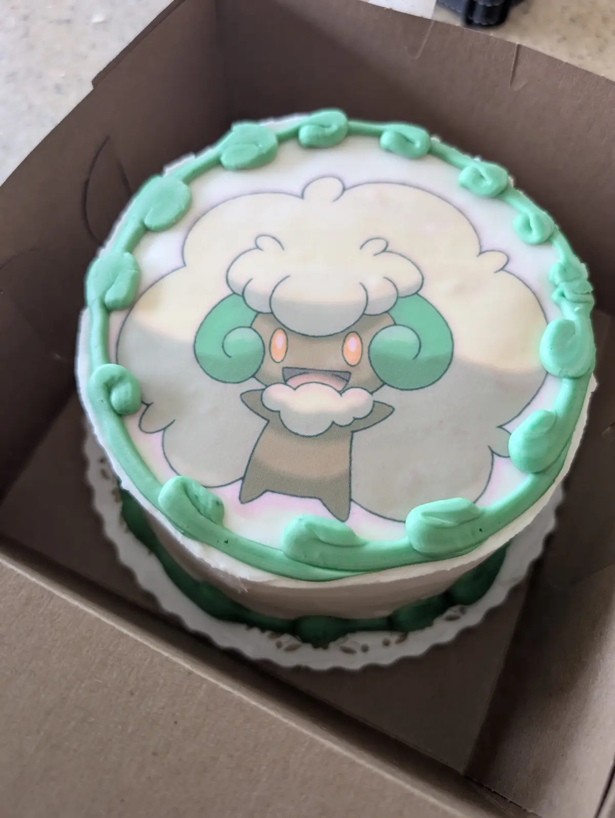 A round cake with white frosting and a green piped border sits in a cardboard box. The top features a printed image of the…