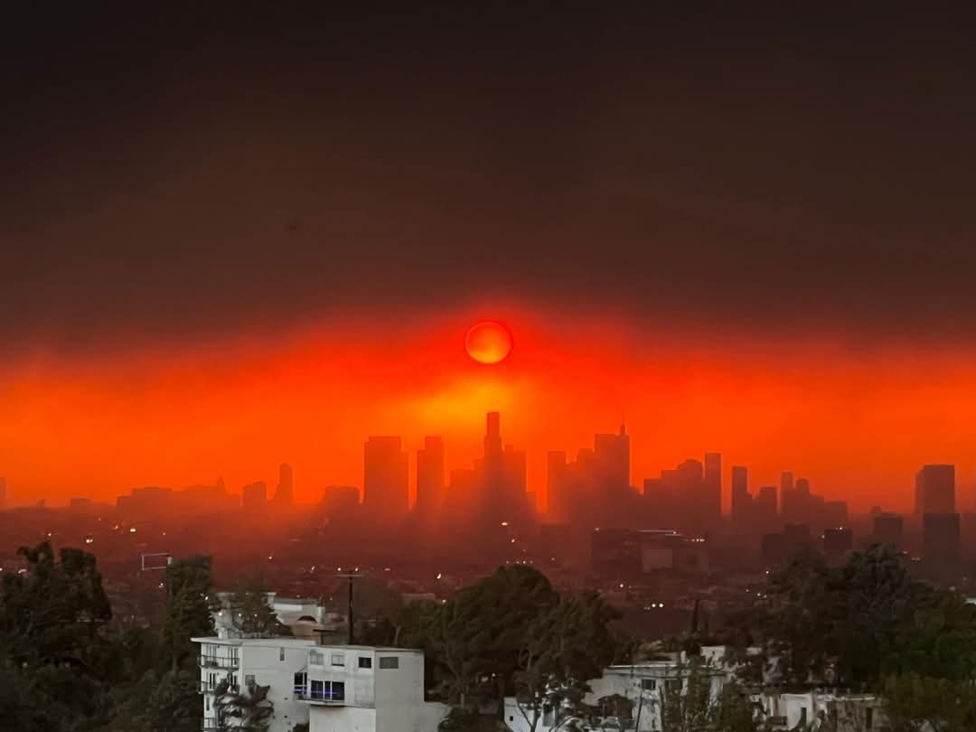 A dramatic, hazy orange sky with a glowing red sun setting over the Los Angeles skyline, silhouetting skyscrapers and…