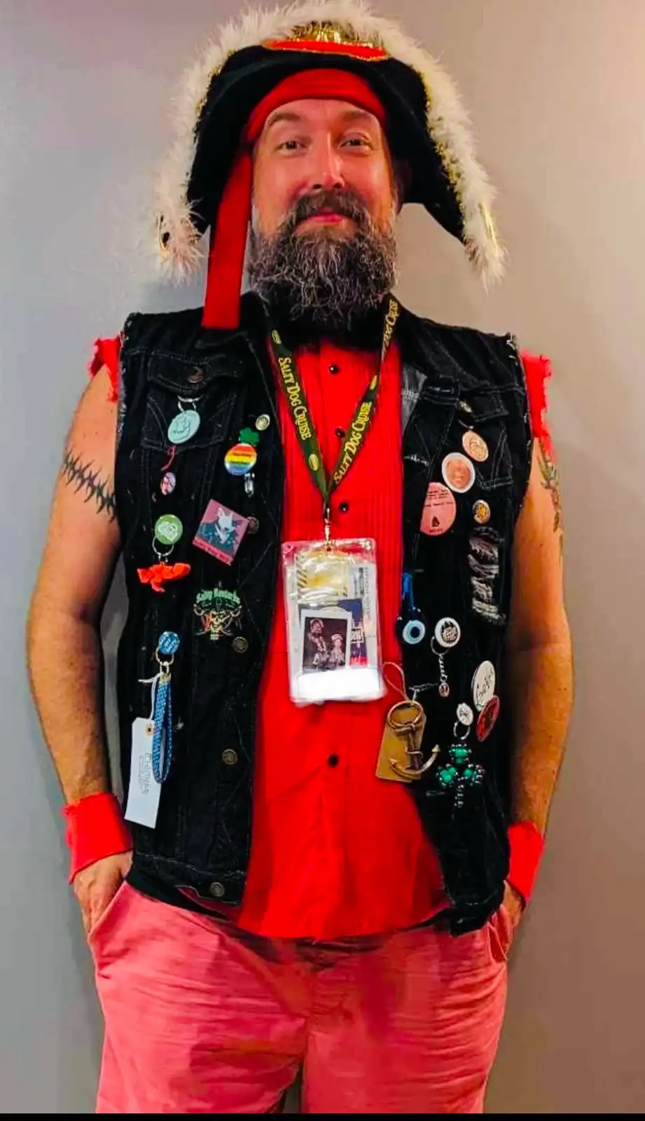 A man with a beard and a black fur-trimmed hat stands against a plain wall, wearing a red shirt, pink pants, and a heavily…