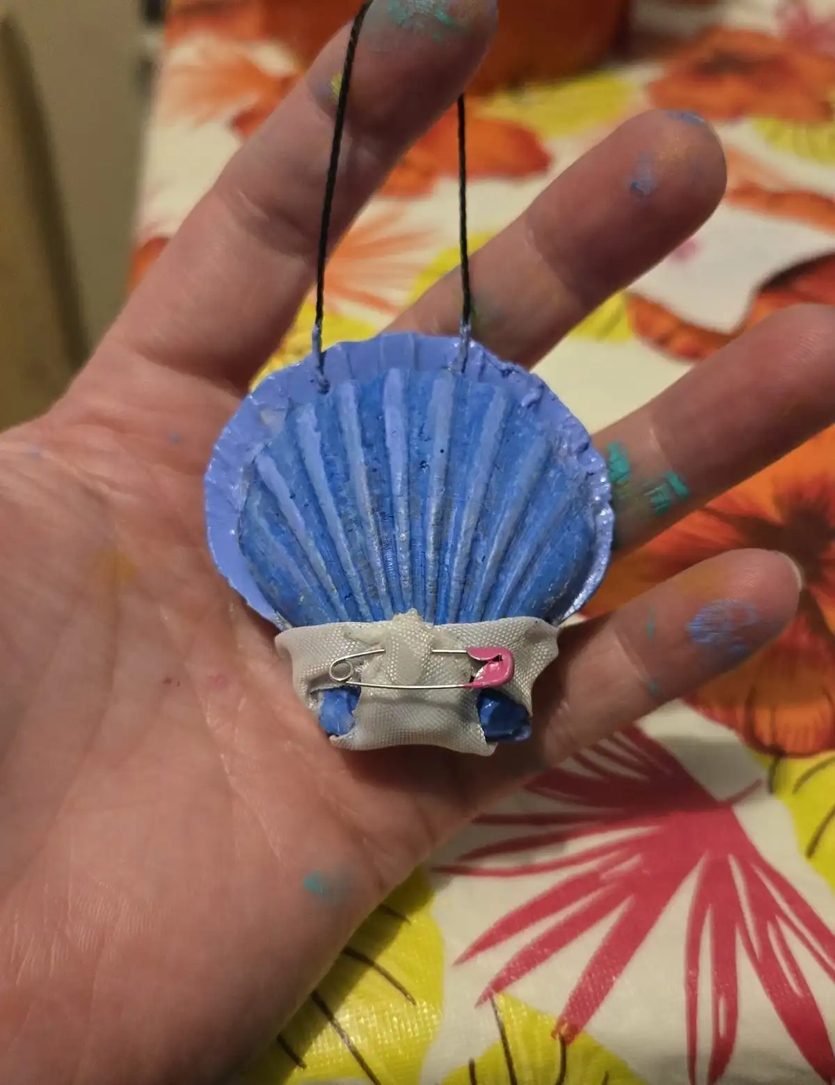 A hand holds a small, blue-painted seashell ornament with black string for hanging. Attached to the shell's base is a piece…