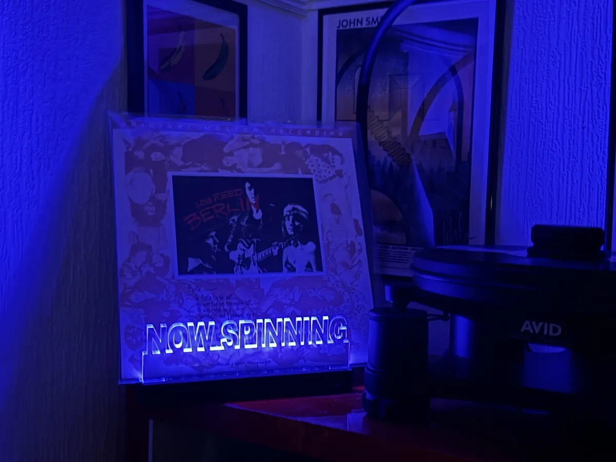 A vinyl record player sits next to a glowing 'NOW SPINNING' sign displaying the album cover for Lou Reed's 'Berlin'. The…