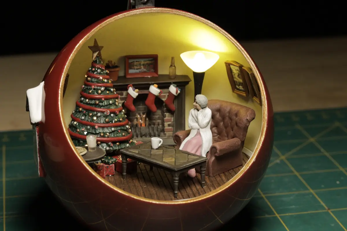 A miniature diorama inside a red Christmas ornament shows an elderly woman in a white robe sitting alone in a brown…