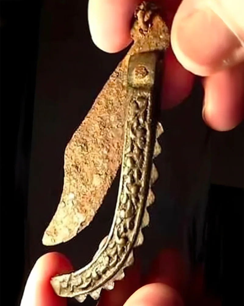 a hand holds an ornate, curved, rusted metal object resembling a scimitar or dagger with intricate carvings along its edge…