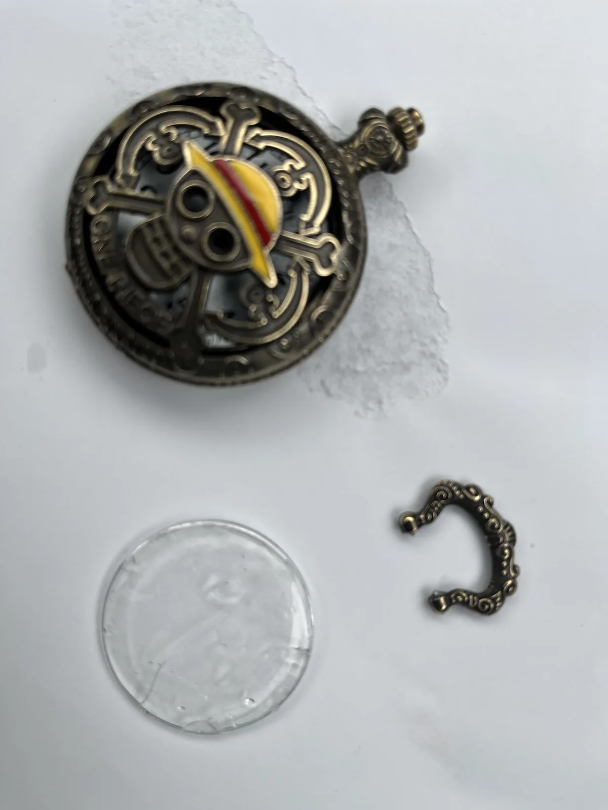 A disassembled pirate-themed pin featuring a yellow hat with a red stripe, set against a skull and crossbones design, lies…