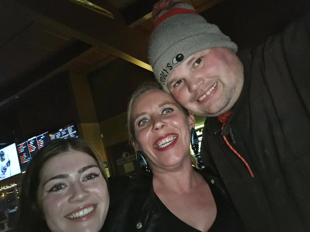 Three people pose for a selfie in a dimly lit bar or restaurant, with a man in a grey beanie with a pom-pom and a logo…