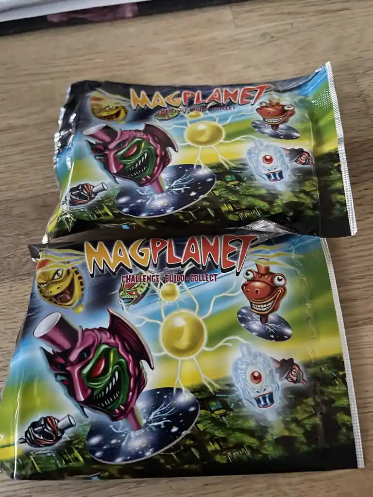Two identical MagPlanet toy packages are stacked on a wooden surface, featuring cartoonish alien heads and the slogan…