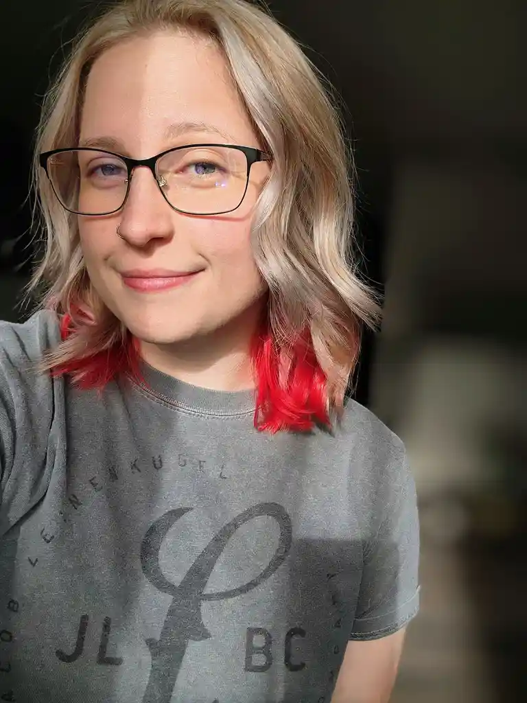 A person with blonde hair and black-rimmed glasses smiles at the camera, wearing a grey t-shirt with a logo. Their hair has…