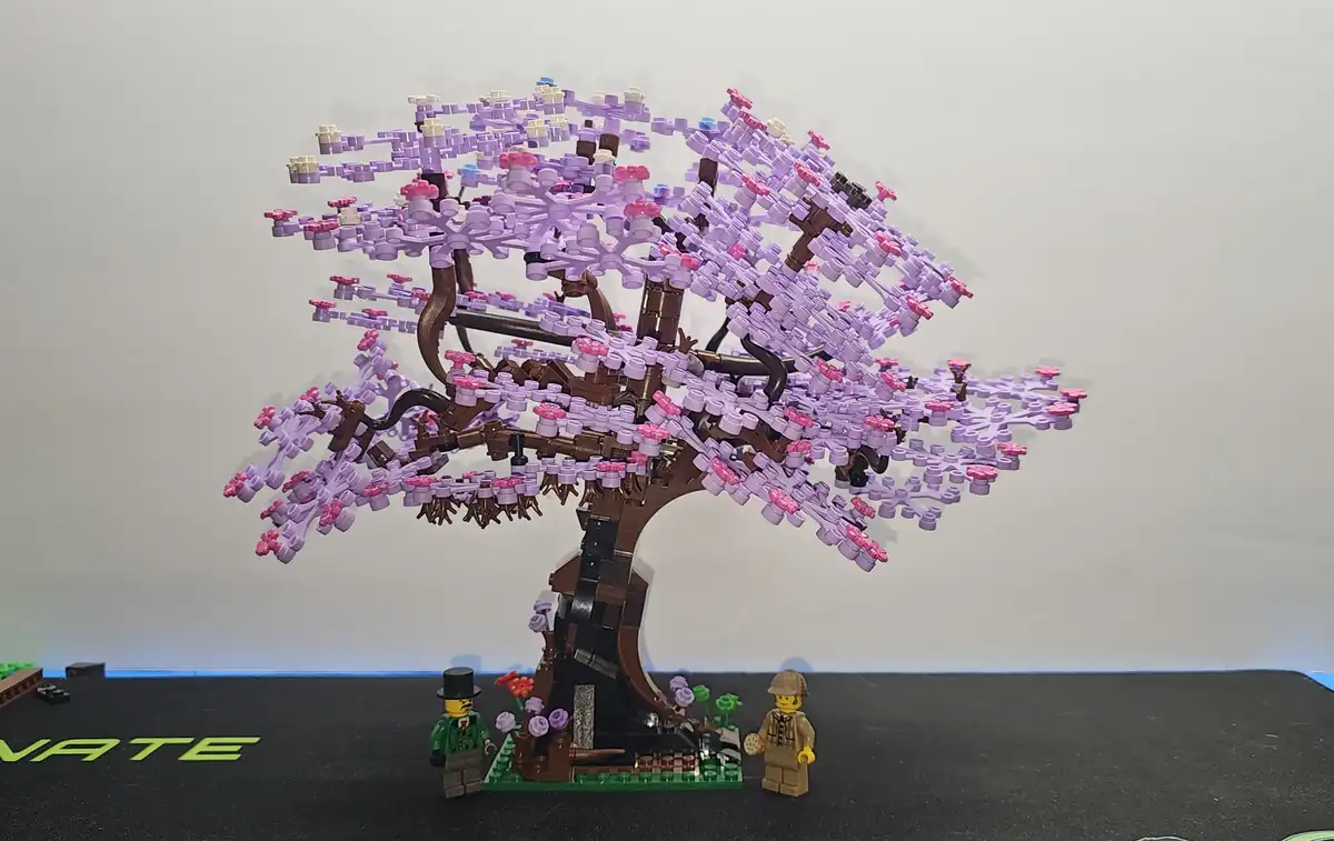 A large, intricate LEGO cherry blossom tree with purple and pink flowers stands against a plain background. Two LEGO…