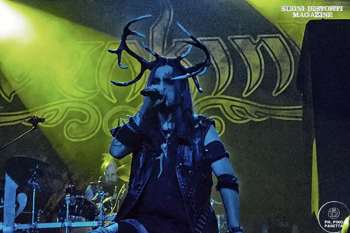 A musician with large antler-like headgear and dark makeup sings into a microphone on a stage lit with green and blue…