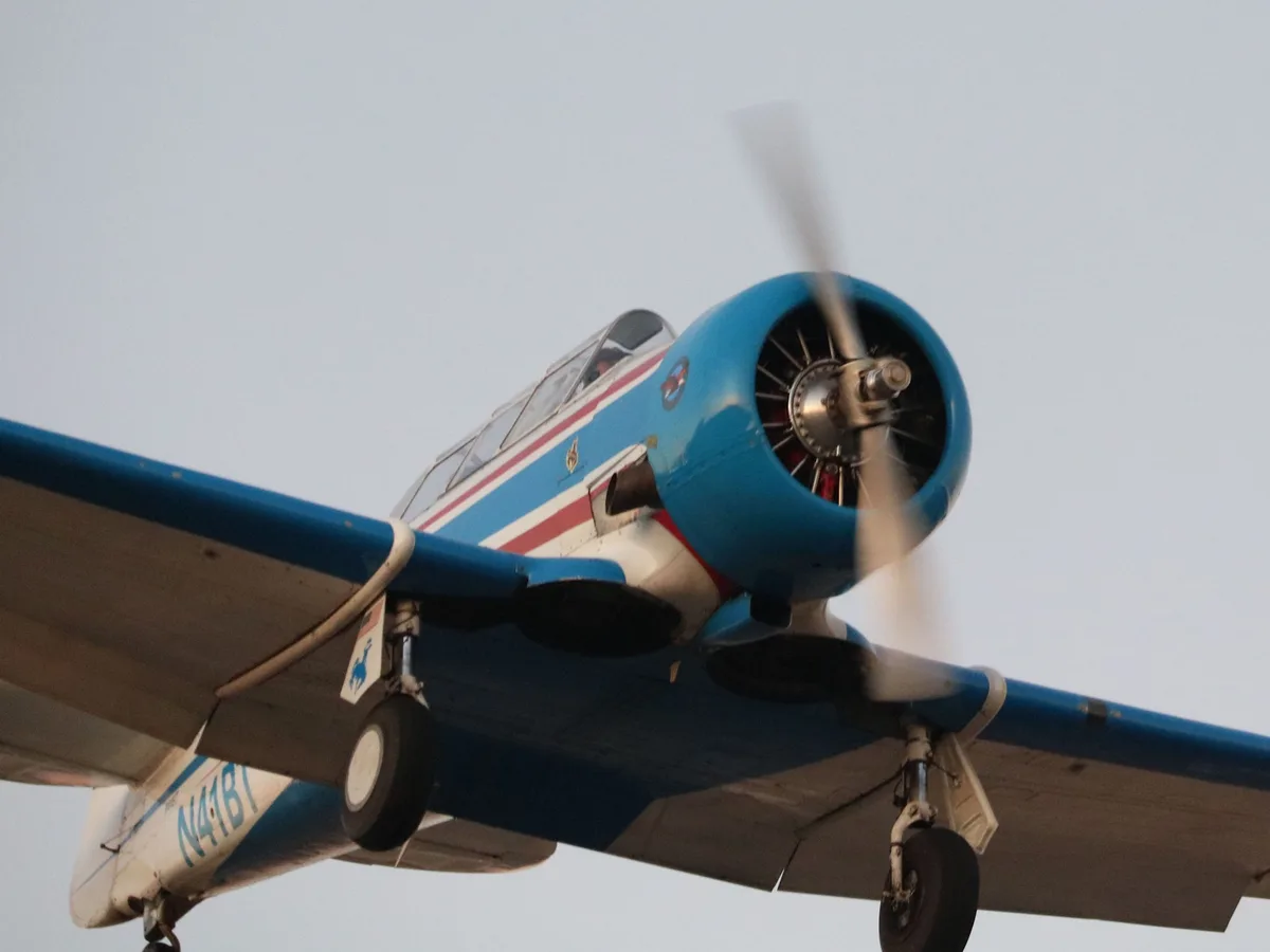 A low-angle shot of a blue, white, and red vintage propeller airplane in flight against a pale sky, with its landing gear…