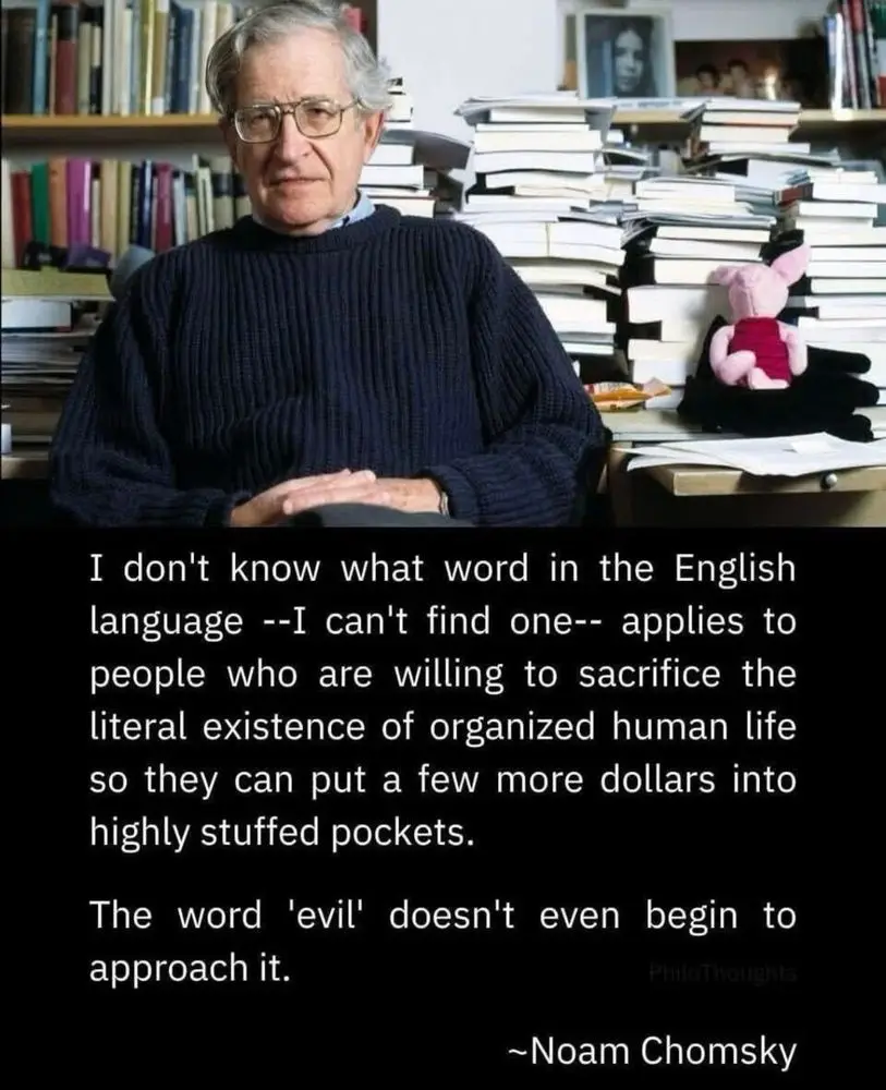 An older man with glasses and a dark sweater sits in front of towering stacks of books and papers. A pink stuffed pig sits…