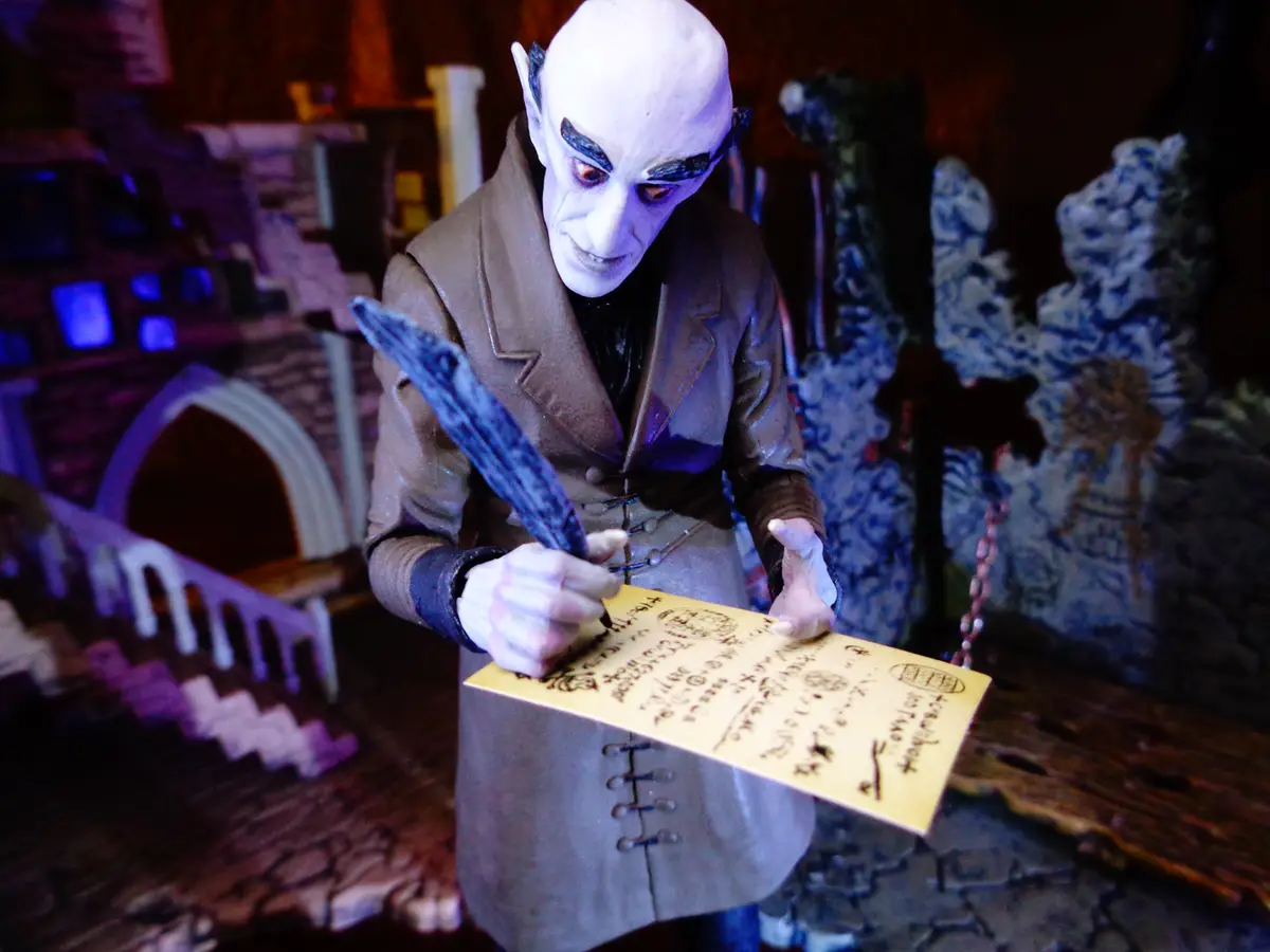 A pale, bald, demonic-looking figurine in a brown coat is writing on a yellow envelope with a quill pen. The background…