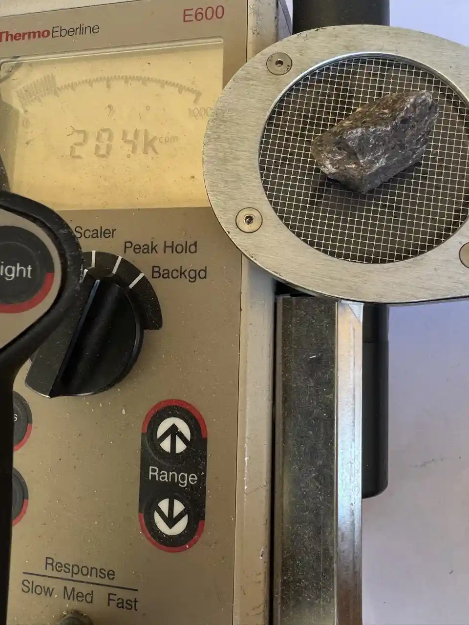 A close-up of a Thermo Eberline E600 radiation detector with a digital display reading '284k cpm' while a rough, dark rock…