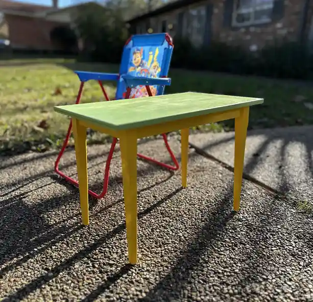 A small, brightly painted wooden desk with a green top and yellow legs sits outdoors on gravel, positioned in front of a…