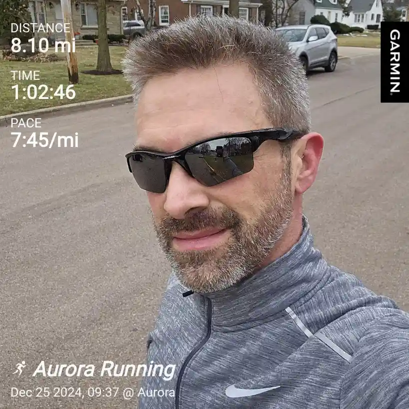 A man in sunglasses and a grey Nike running jacket takes a selfie outdoors on a residential street, with a Garmin fitness…