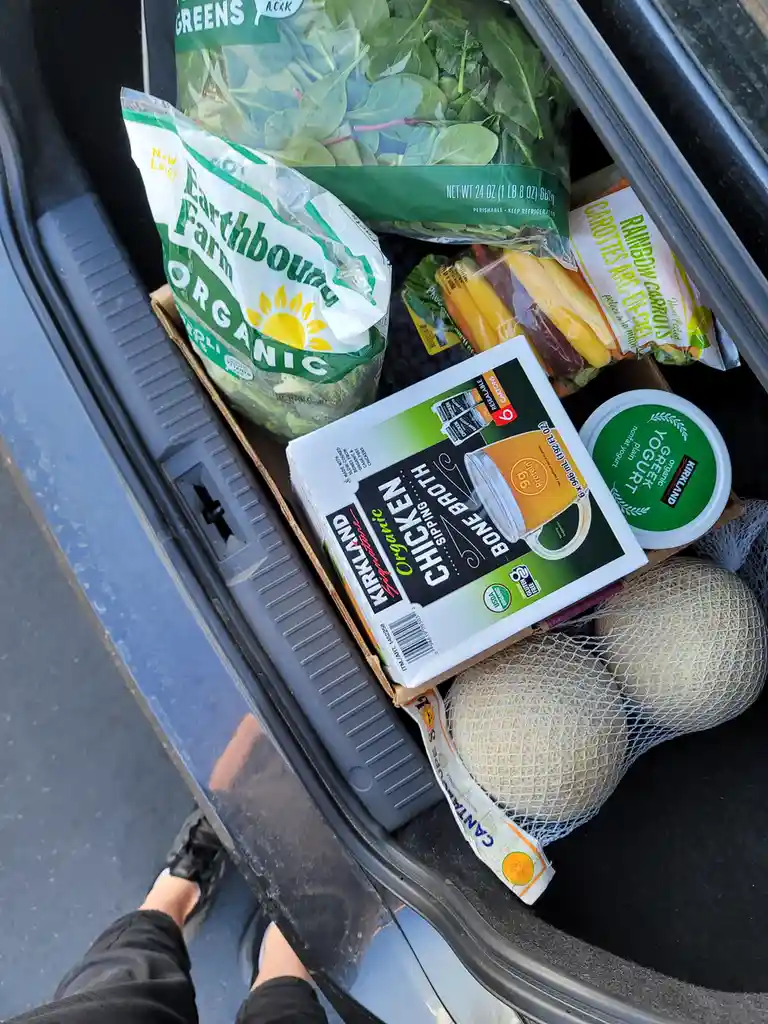 An overhead view of a car trunk filled with groceries including Earthbound Farm organic greens, Kirkland organic chicken…