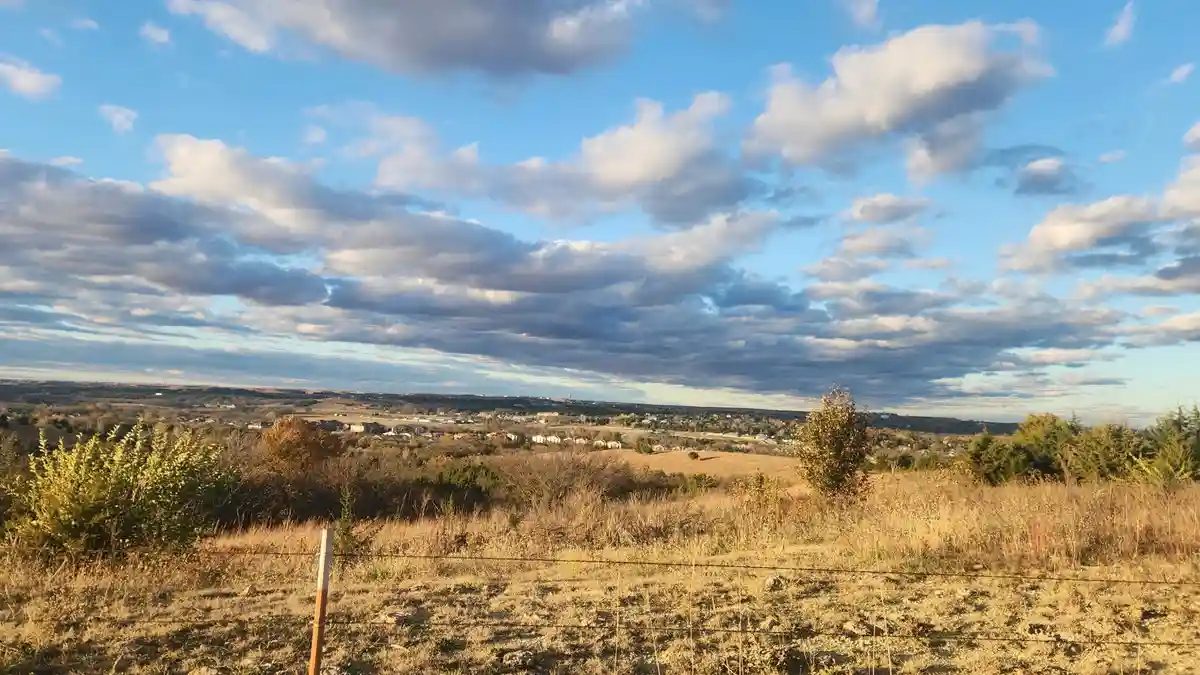 A scenic landscape photo shows a wide field with dry grass and scattered bushes under a partly cloudy blue sky, with a…