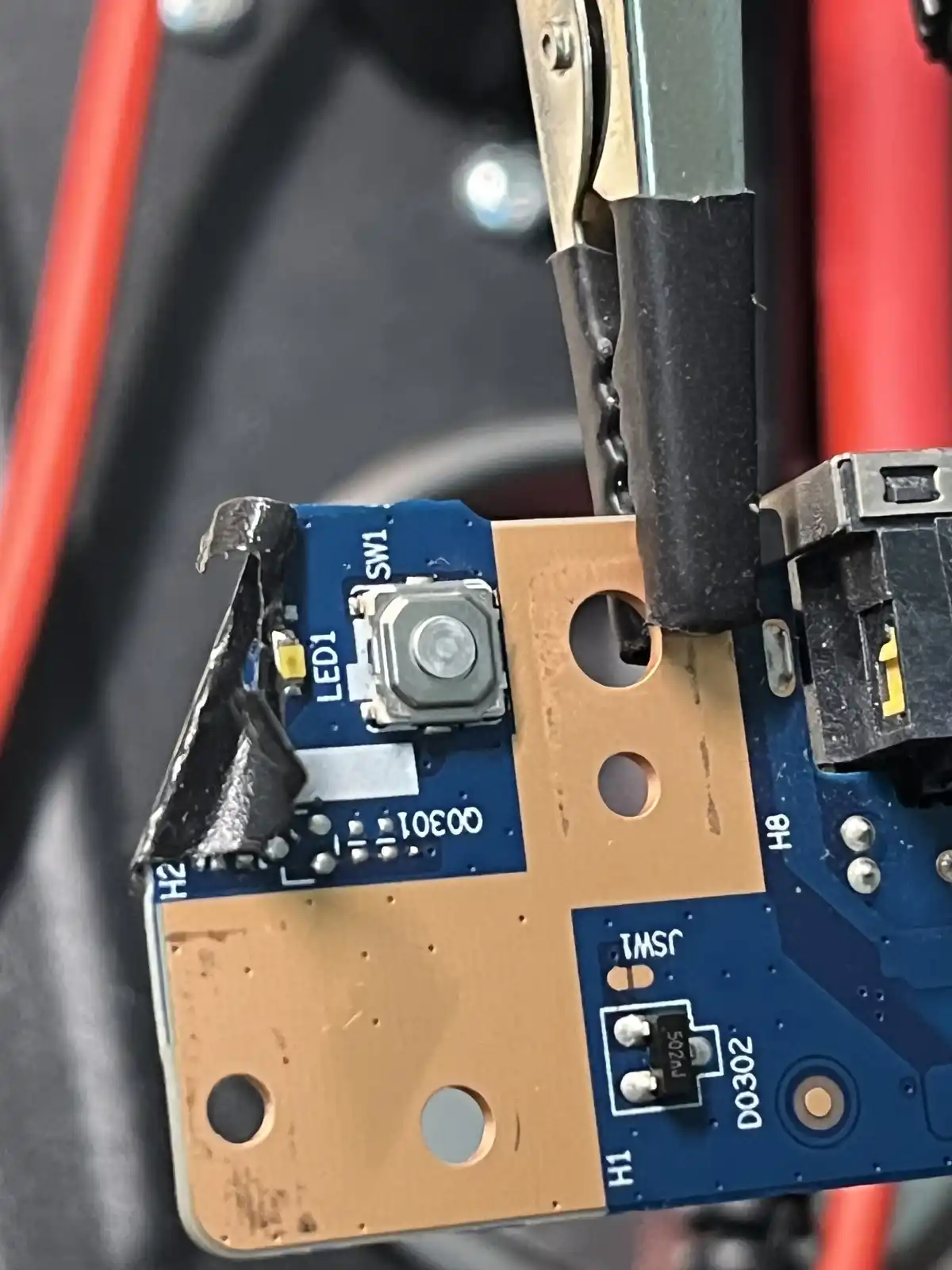 A close-up photo shows a damaged blue and copper circuit board with a torn piece of black tape, a small button labeled…