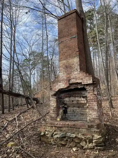 A dilapidated brick fireplace stands in a forest with bare trees and fallen leaves under a partly cloudy sky. The structure…
