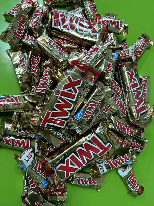 A large pile of individually wrapped Twix chocolate bars with gold foil packaging and red 'Twix' branding is scattered…