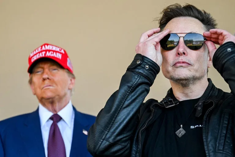 elon musk, wearing sunglasses and a black leather jacket, adjusts his glasses while looking forward, with donald trump in…