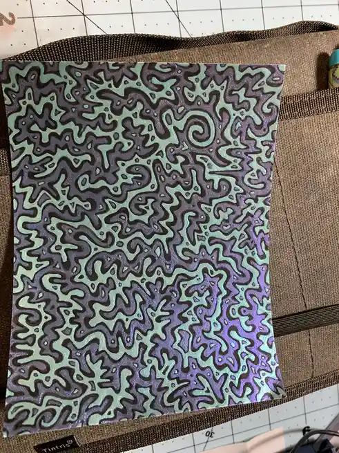 A handmade, glittery, psychedelic patterned paper with swirling, organic shapes in mint green and purple hues, resting…