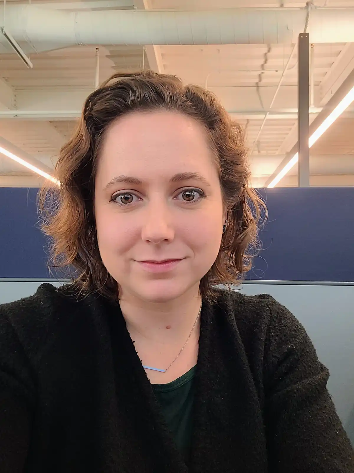 A woman with short, wavy brown hair and a subtle smile looks directly at the camera in an office setting with visible…