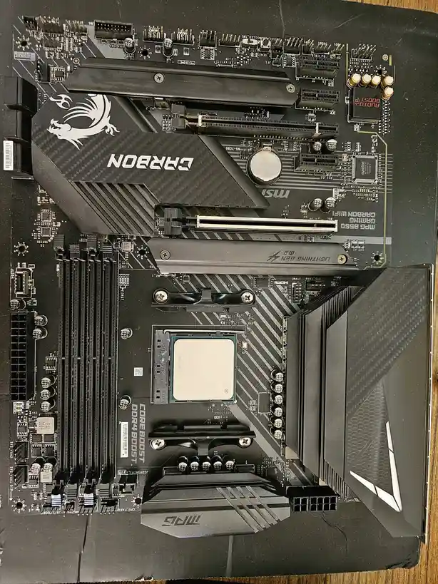 A top-down photo of a black MSI MAG motherboard with a white AMD Ryzen 7 5800X3D CPU installed in the socket. The board…