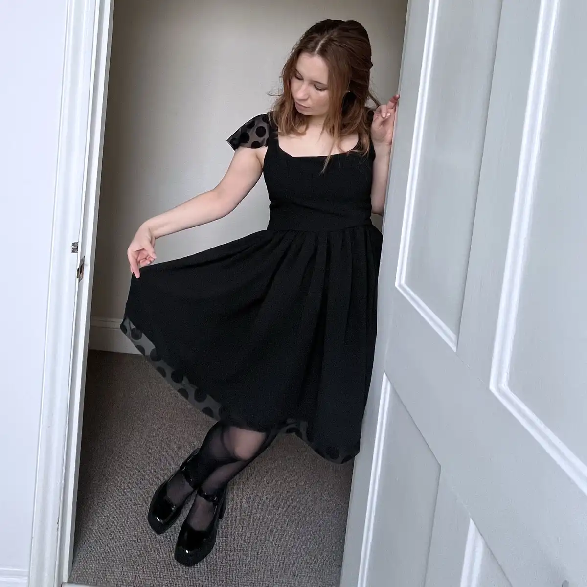 A woman with brown hair stands in a doorway wearing a black dress with sheer polka-dot sleeves and hem, black tights, and…
