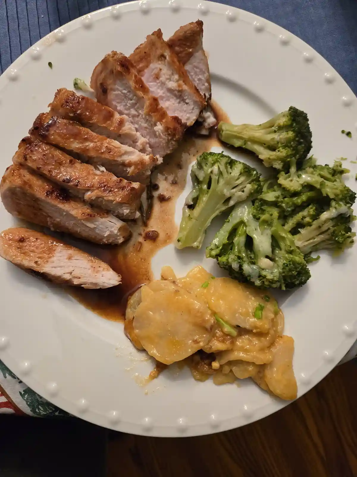 A white plate holds sliced grilled chicken breast with a brown sauce, steamed broccoli with cheese, and cheesy potato…