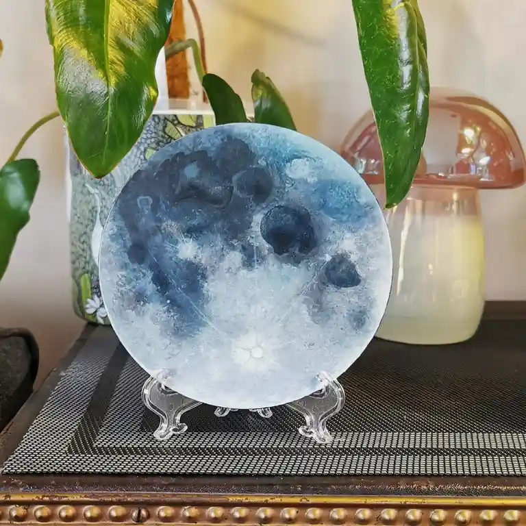 A decorative, circular blue moon sculpture with crater-like textures sits on a clear stand atop a patterned mat. It's…