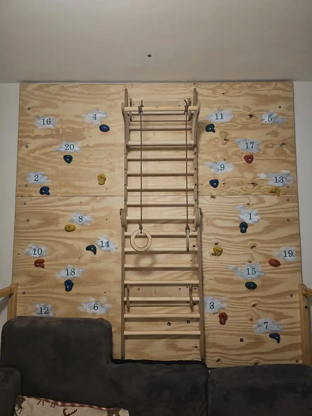 A wooden indoor climbing wall with colorful handholds and painted cloud shapes containing numbers 1-20, flanked by a wooden…