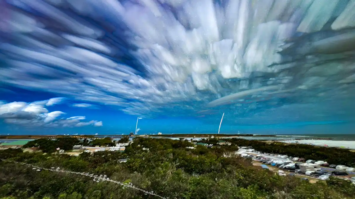A long-exposure photograph shows a dramatic, streaked sky over a coastal landscape with trees, houses, and parked vehicles…