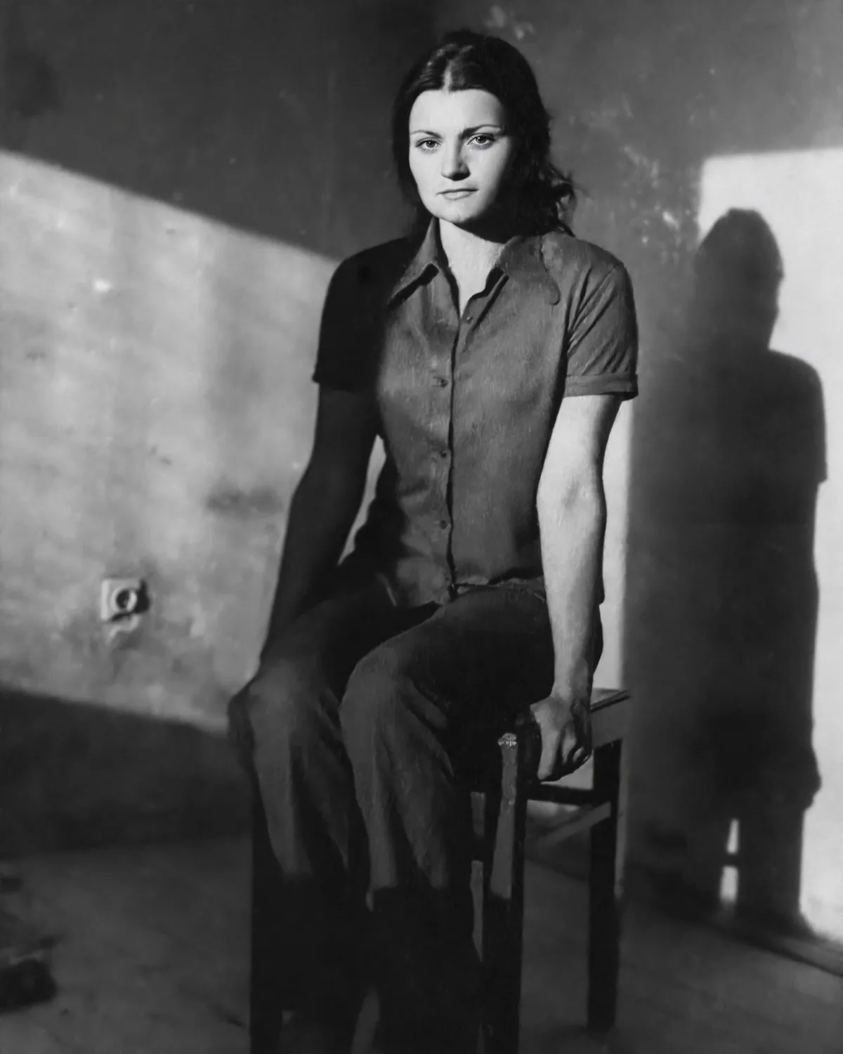 A black-and-white photograph shows a woman sitting on a wooden chair in a dimly lit room with a stark shadow cast on the…