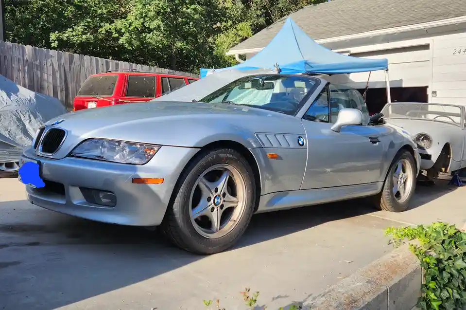 A silver BMW Z3 roadster is parked in a driveway under a blue pop-up canopy, with a red SUV and a covered car behind it. The…