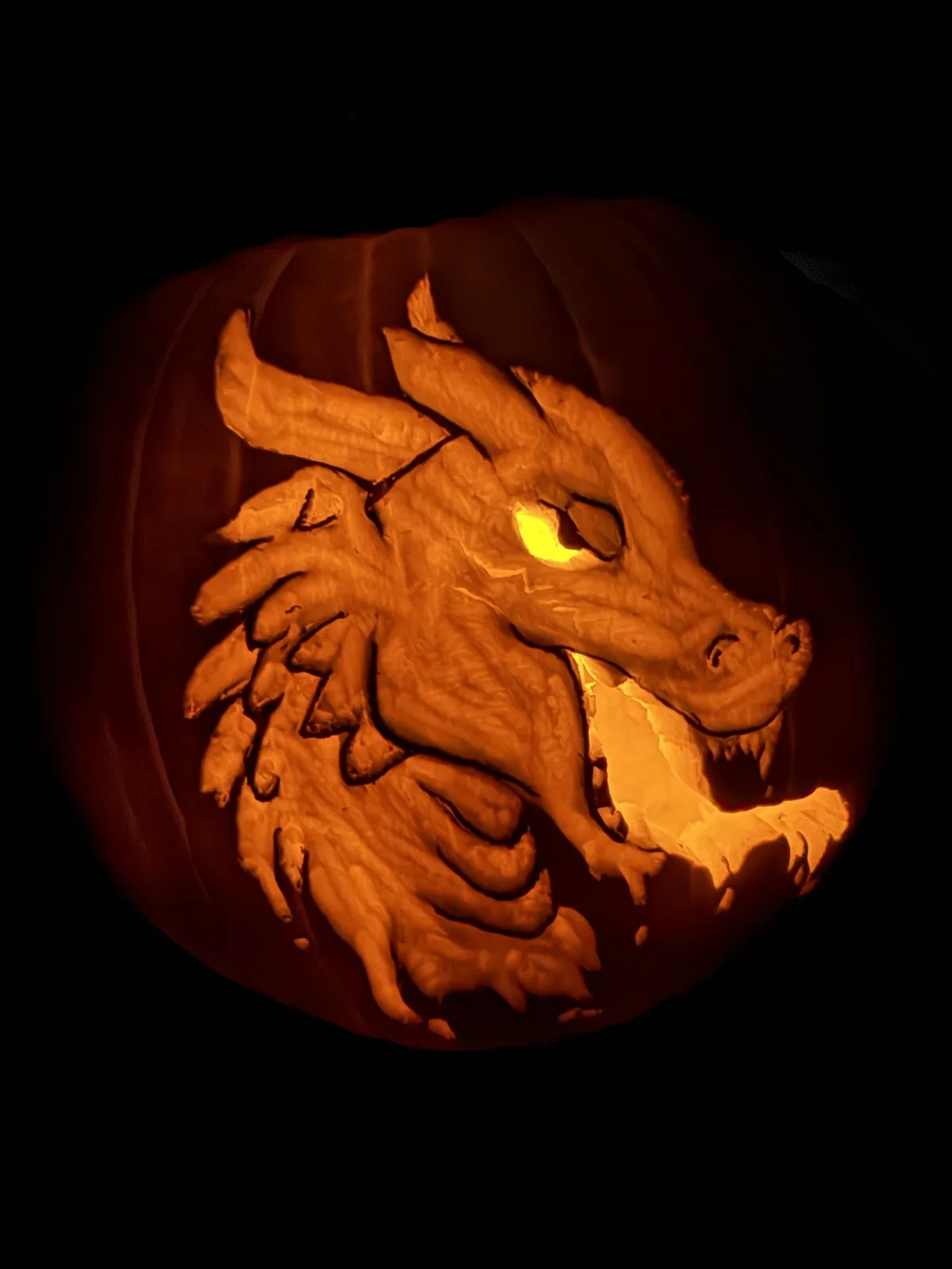A carved pumpkin illuminated from within, featuring a detailed dragon design with wings, scales, and an open mouth, set…