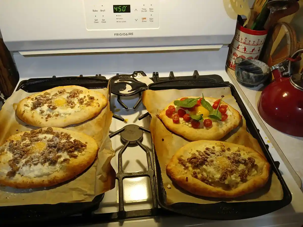 Four small flatbreads with raw eggs, crumbled meat, and cheese are baking on two trays on a stovetop. One pizza has cherry…