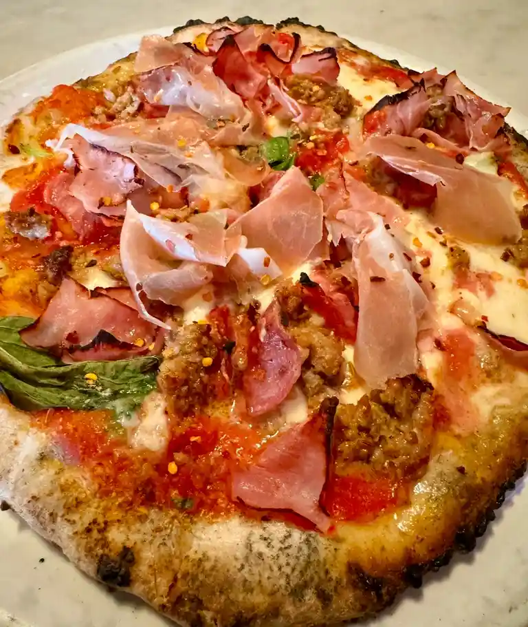 A close-up photo of a rustic pizza with a charred crust, topped with melted cheese, tomato sauce, crumbled sausage, and thin…