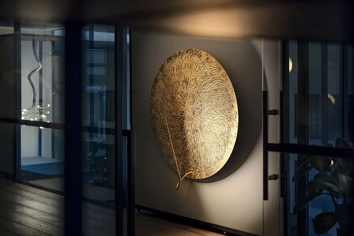 A large, circular, textured golden wall art piece is mounted on a white wall in a modern interior with glass partitions and…