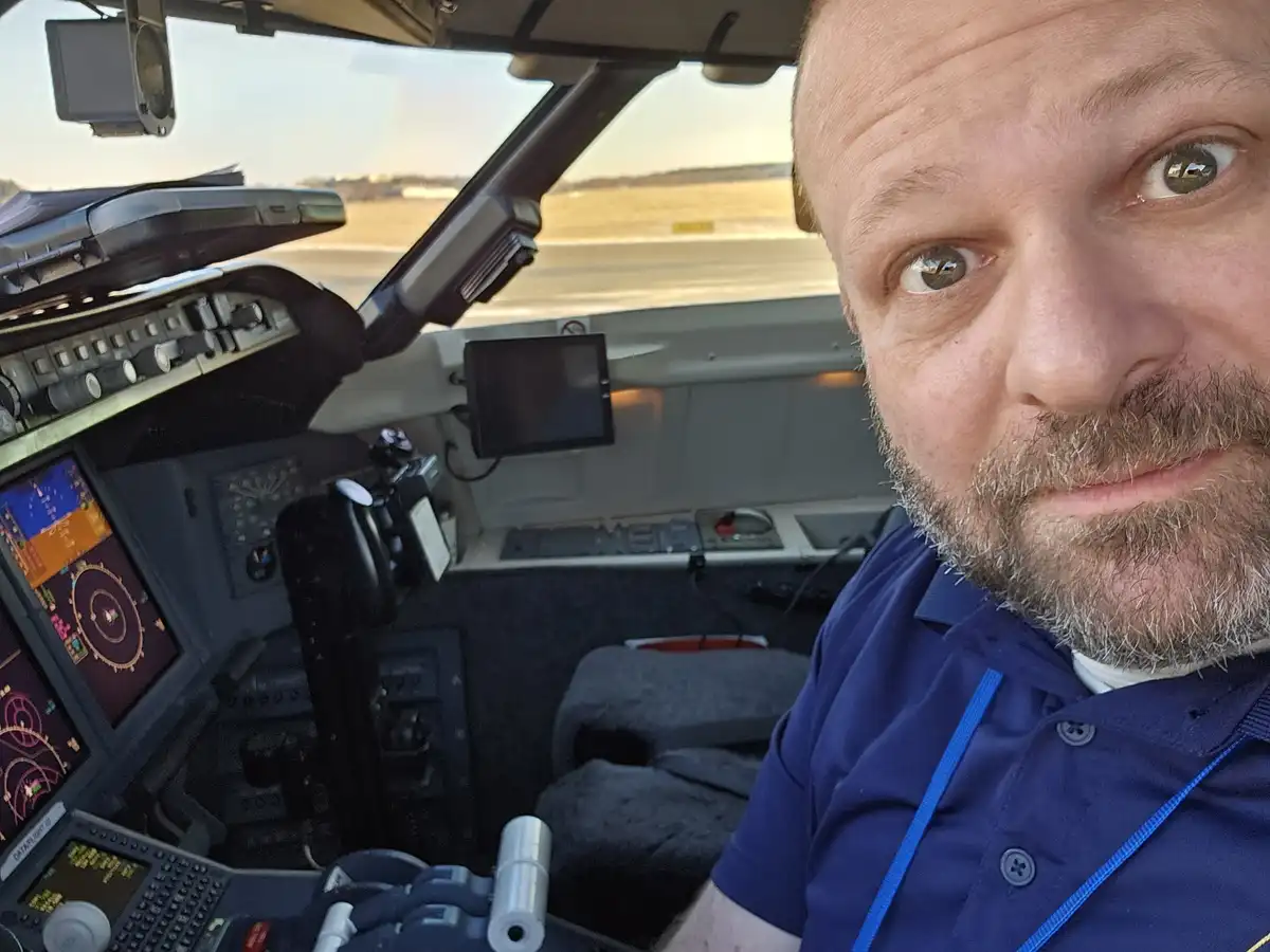 A man with a beard and blue shirt takes a selfie from inside an airplane cockpit, looking directly at the camera with wide…