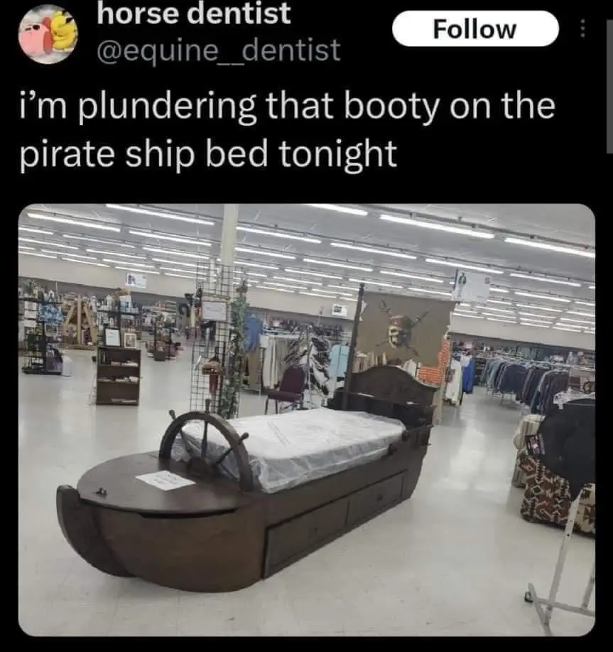 A dark brown wooden bed shaped like a pirate ship with a steering wheel at the headboard and a mattress on top, displayed in…