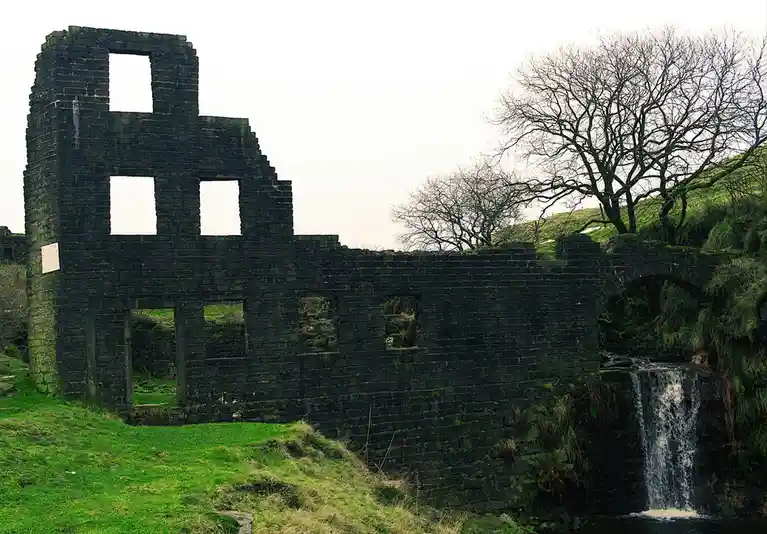 The image shows the dark stone ruins of a building with multiple window and door openings, set against a bright sky. A small…