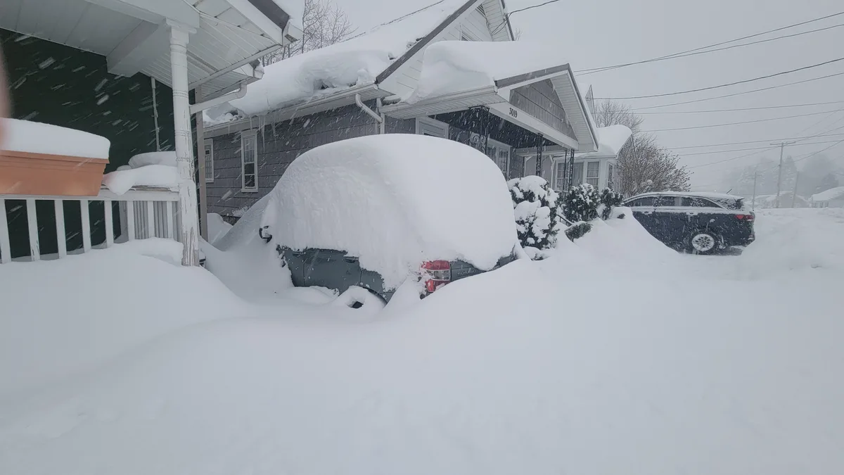 A heavy snowfall blankets a residential street, burying a gray Subaru almost entirely under deep snow, with only its rear…