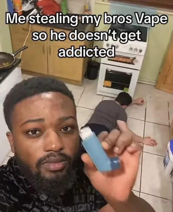 A man with a beard holds up a blue inhaler while looking at the camera, with another person lying motionless on the kitchen…