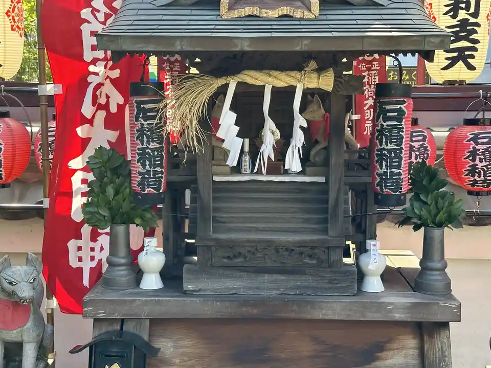 A small Japanese shrine with a thatched roof, decorated with red lanterns bearing the text '石橋稲荷', white paper streamers…