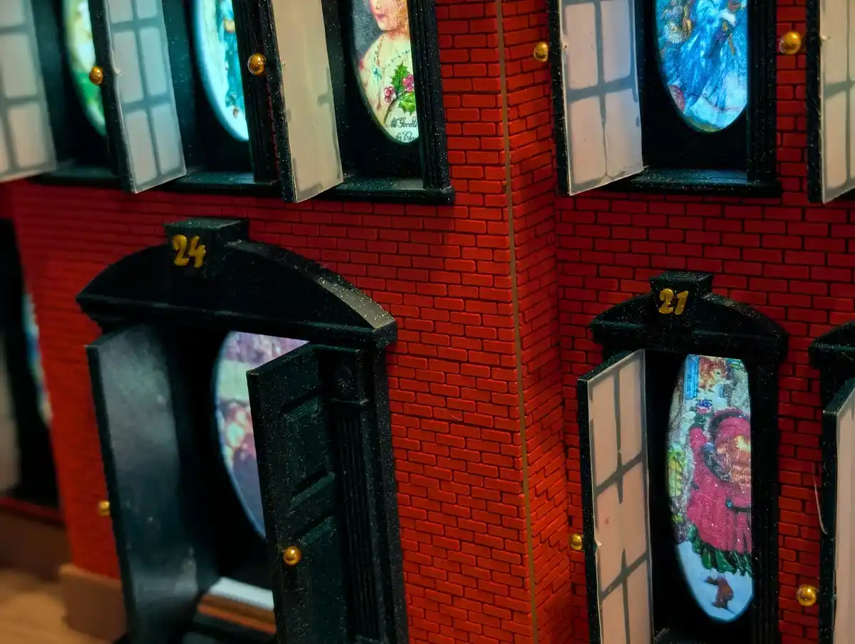 A close-up of a red brick miniature building with black doors and windows, each displaying a different colorful, painted…