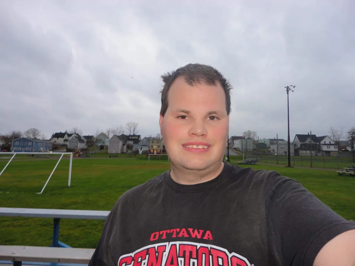 A man in a dark grey Ottawa Senators t-shirt takes a selfie on a cloudy day at a sports field with a soccer goal and…